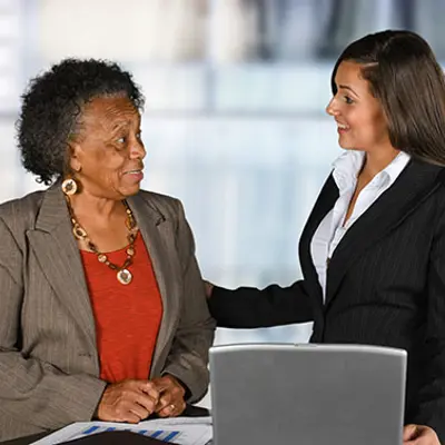 a younger woman in professional dress speaking to an older woman standing at a table with papers and a laptop