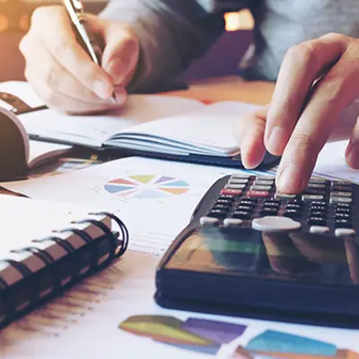 closeup of someone sitting at a table filled with papers, holding a pen, writing in a notebook while using a calculator