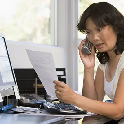 a woman on a phone sitting at a desk in front of a computer holding a piece of paper looking concerned
