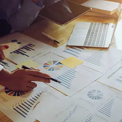 closeup of the hands of two people at a table pointing at papers with charts and tables and using a laptop