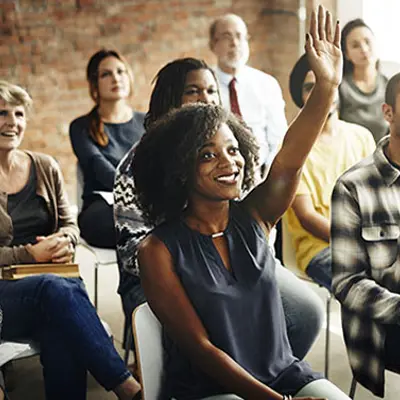 a diverse group of people sitting in a room, in the foreground a woman is raising her hand to ask a question and smiling