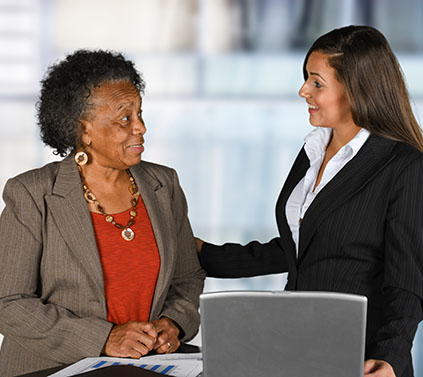 a younger woman in professional dress speaking to an older woman standing at a table with papers and a laptop