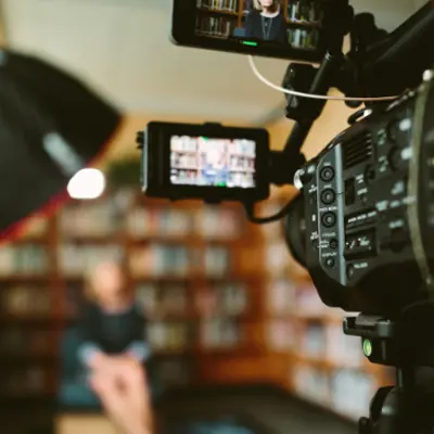 a video camera and a light in the foreground with someone sitting and being filmed in front of a bookcase in the background