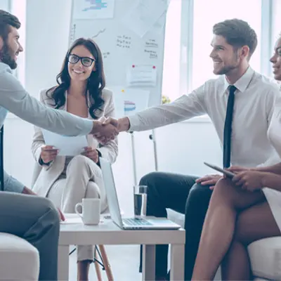 a group of four people sitting together in an office, two men are shaking hands while two women look on