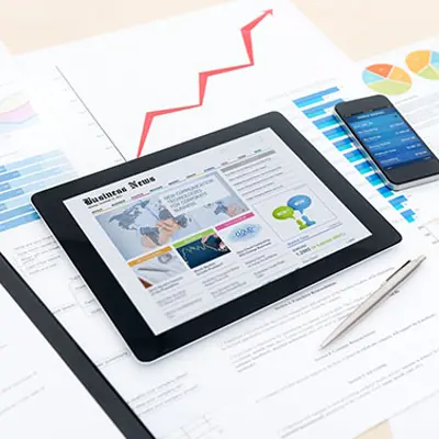 a tablet with news on the screen, on a table filled with papers, a pen and a phone