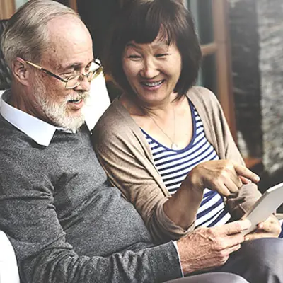 a middle-aged couple sitting together, the woman is smiling and pointing at a tablet that the man is holding