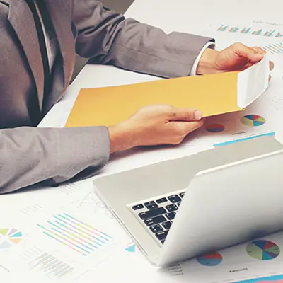 closeup of someone holding a manila envelope over a table filled with paperwork next to a laptop