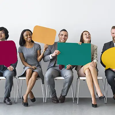 a group of six diverse men and women sitting on chairs facing the camera, smiling while holding large colourful speech bubbles